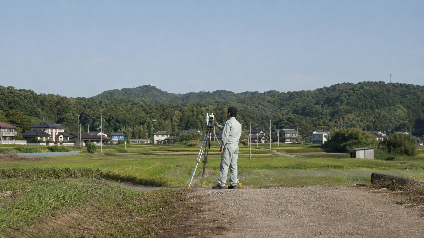郡山市郊外の里山と田畑を背景に、トータルステーションで測量する土地家屋調査士